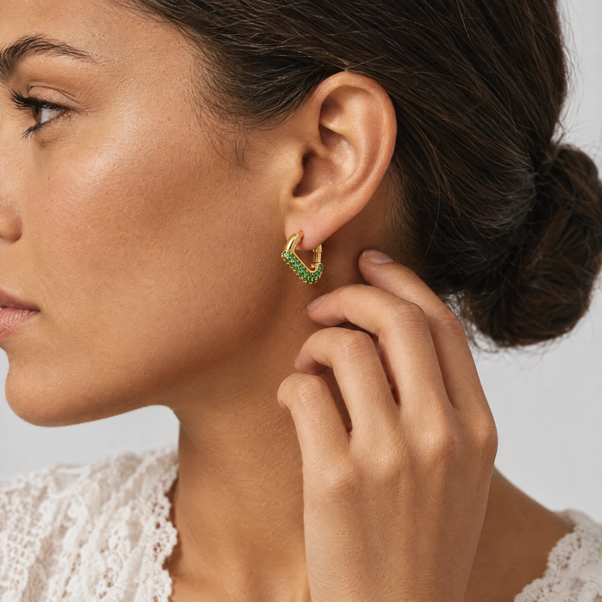 Woman touching her ear while wearing a gold Cora Crystal Heart Hoop Earring with green crystals