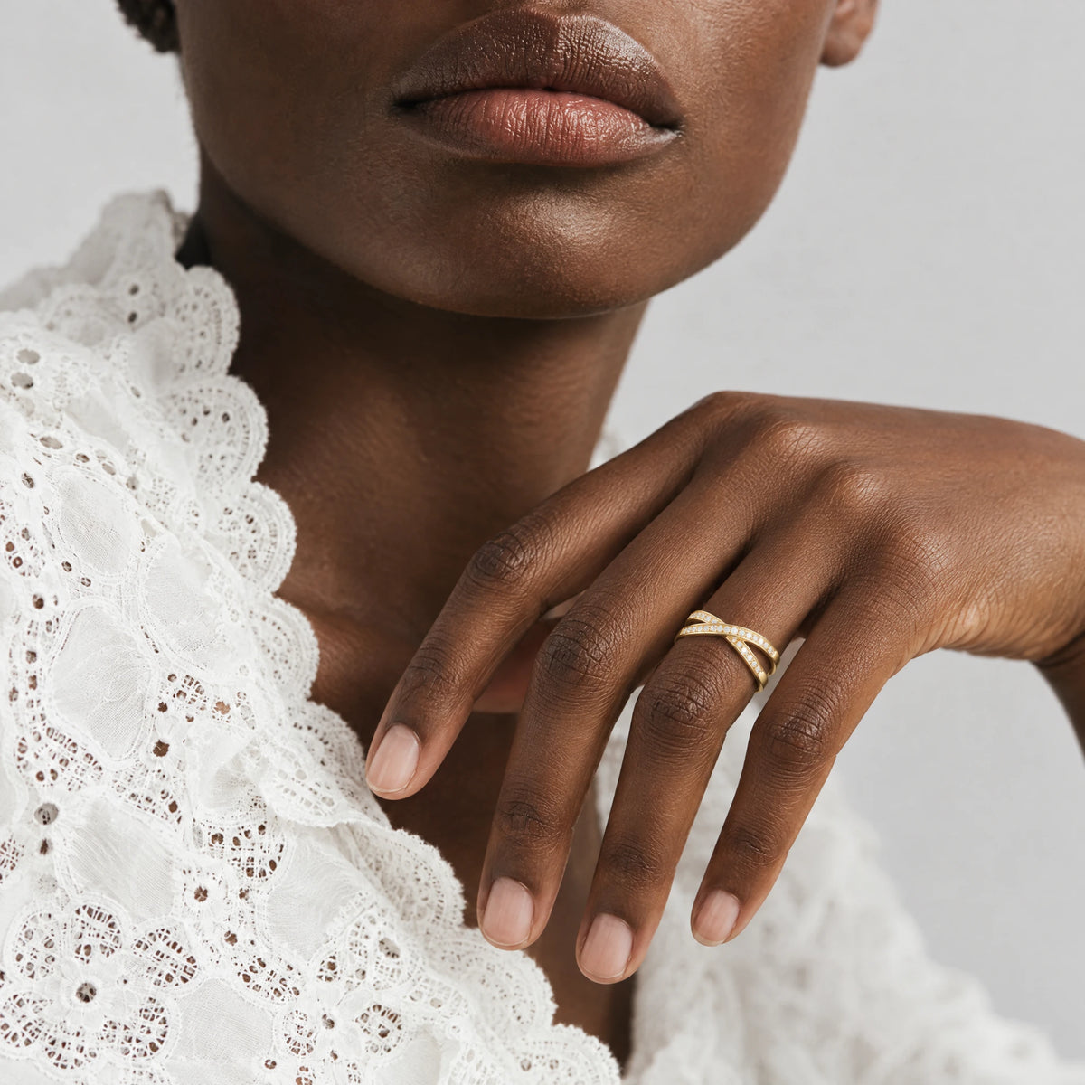 Woman's hand near her collarbone wearing the gold Ivy Crossover Crystal Ring, white lace blouse