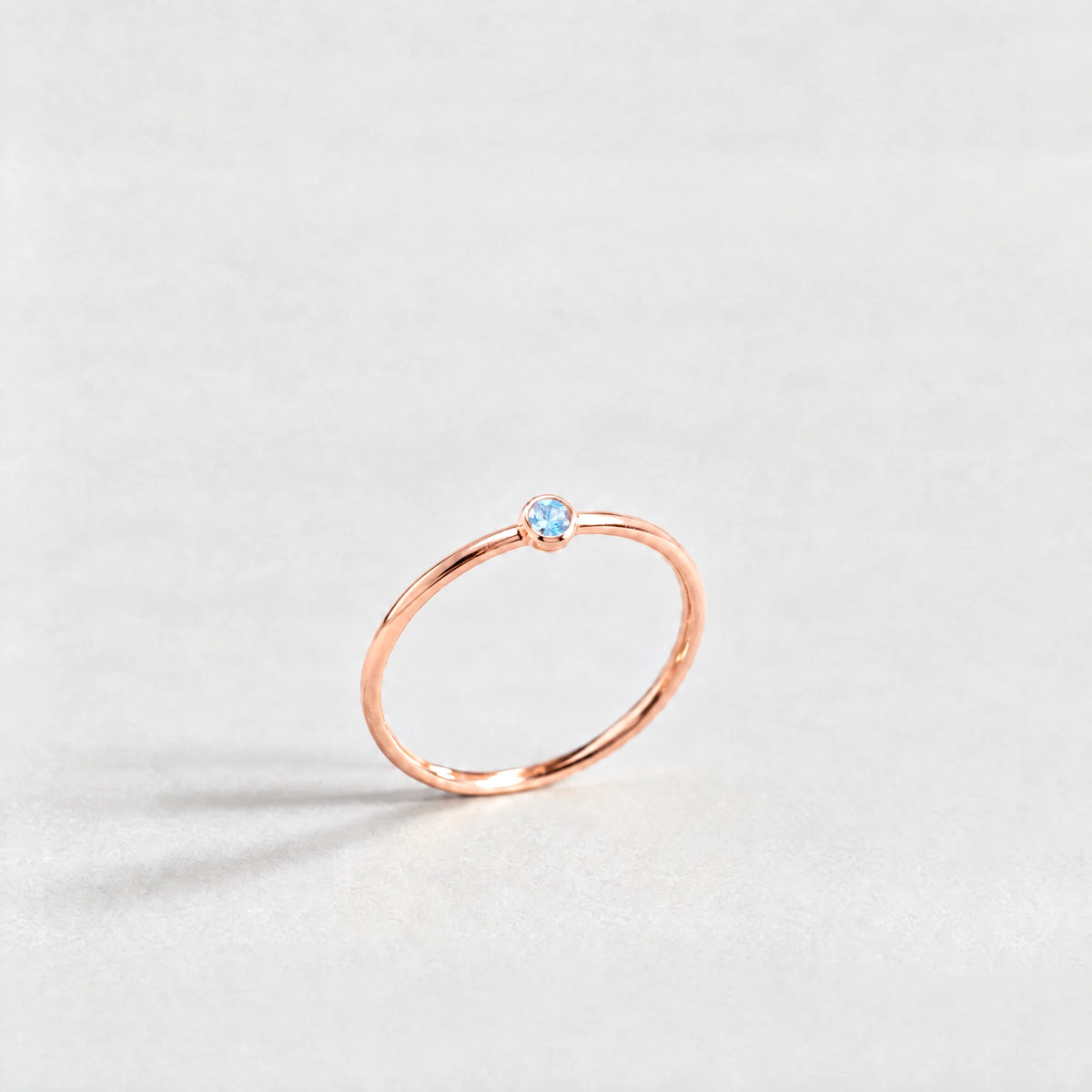 Rose gold Lena Birthstone Ring with aquamarine blue March birthstone-color crystal on a light grey background