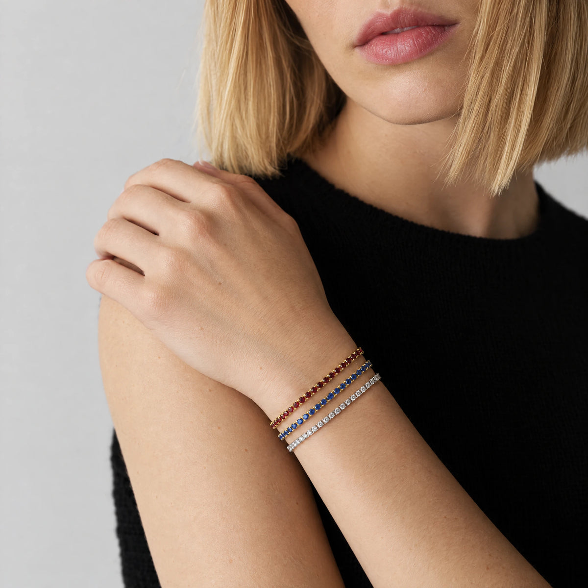 Woman's hand resting on her shoulder, wearing a stack of three delicate MAYA Birthstone Tennis Bracelets in garnet red, sapphire blue and clear crystals