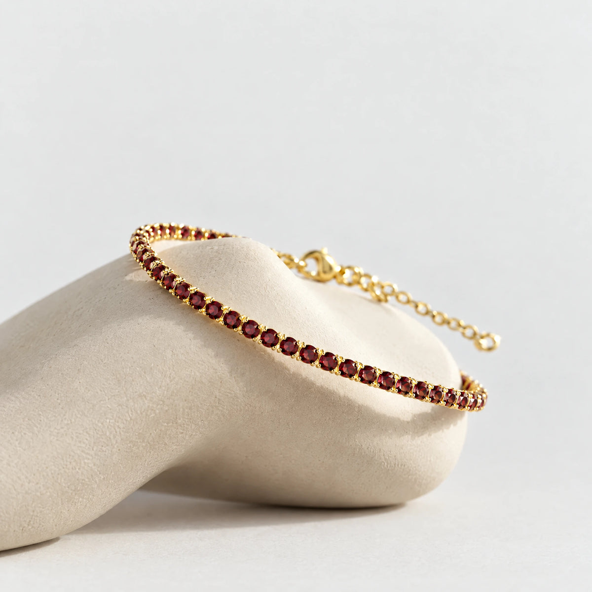 Gold MAYA Birthstone Tennis Bracelet with garnet-red crystals resting on a cream clay pedestal
