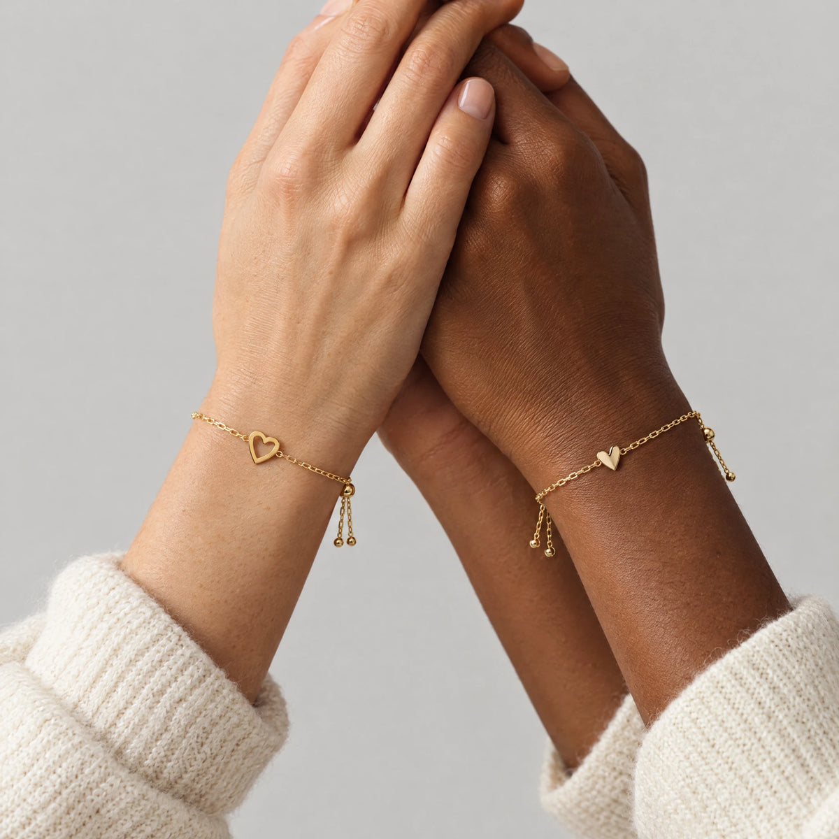 Two friends holding hands, each wearing one bracelet from the gold Rosie Two Hearts Bracelet Set
