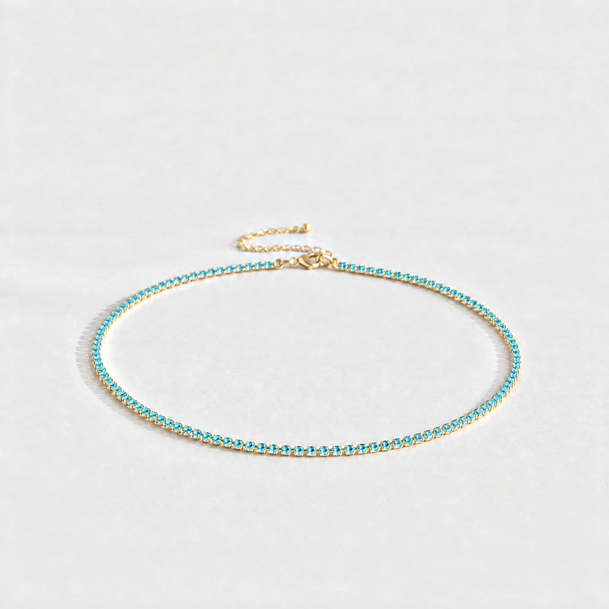 Gold Tessa Birthstone Tennis Necklace with topaz blue December birthstone-color crystals on a light grey background