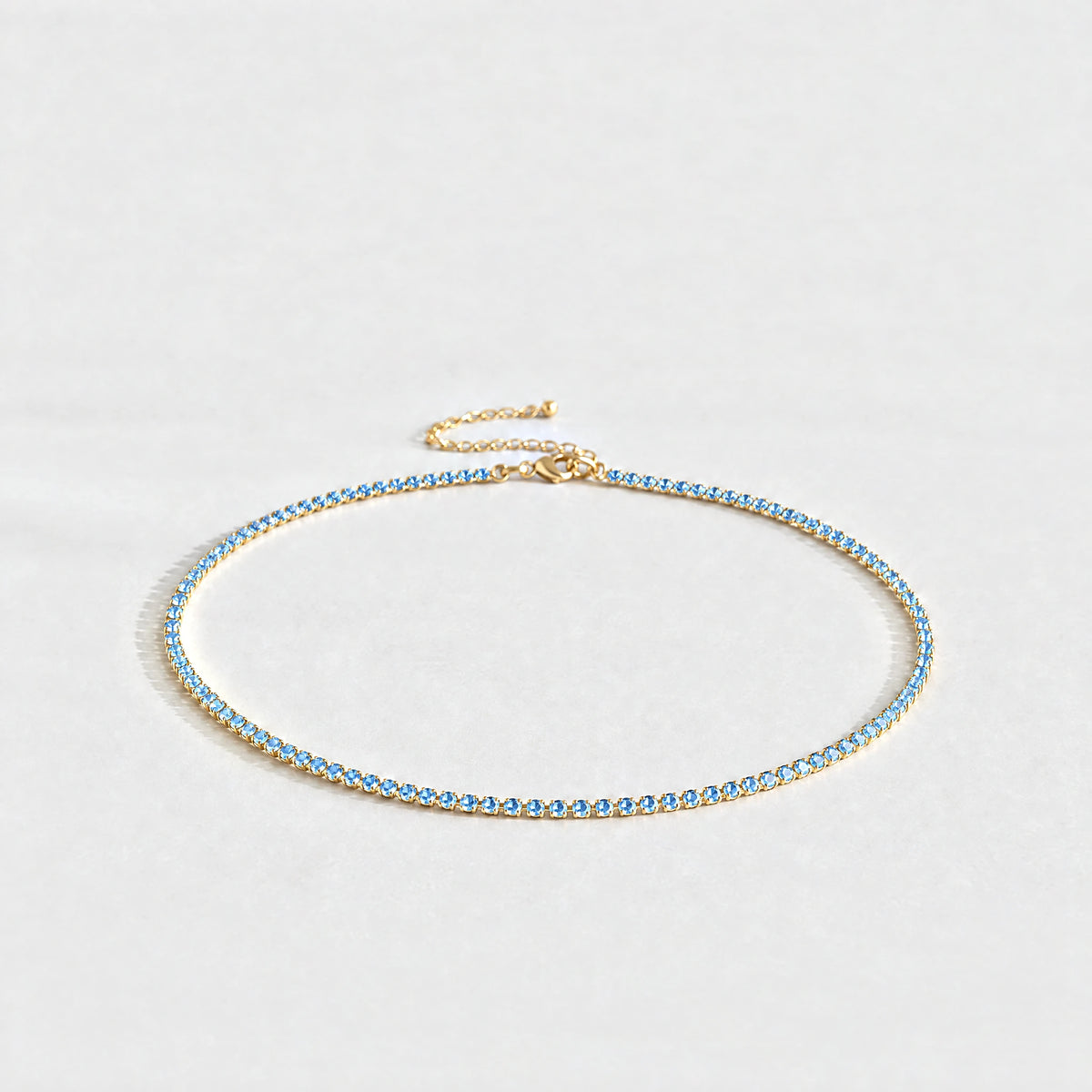 Gold Tessa Birthstone Tennis Necklace with aquamarine blue March birthstone-color crystals on a light grey background